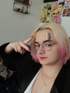 Alex, a white femme person with pink dip-dyed blonde hair, wearing face paint of the bisexual and non-binary flags